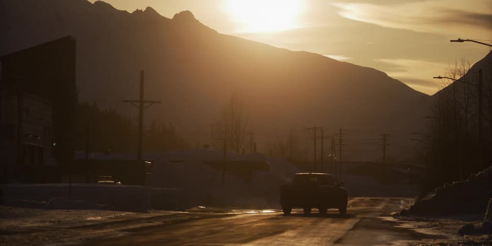 A pickup truck and an SUV are silhouetted as they drive down a snowy street in a mountain town during a beautiful golden hour sunset