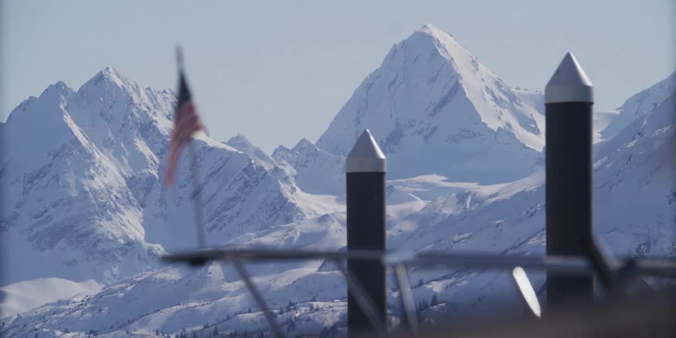 A majestic snow-covered mountain range rises in the background behind an out-of-focus American flag