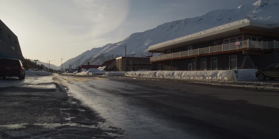 A slow tracking shot reveals a quiet, snow-lined street in a remote mountain town during a cold winter afternoon