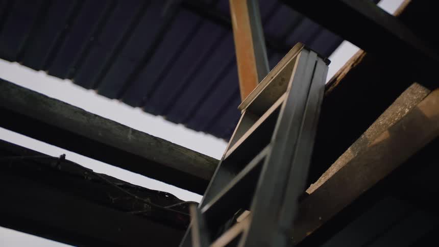 Rain falls on a metal ladder resting against wooden beams under a corrugated roof