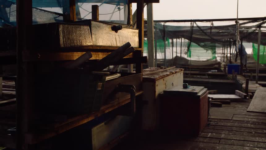 A fisherman works on a rustic floating fish farm during a quiet, hazy sunrise