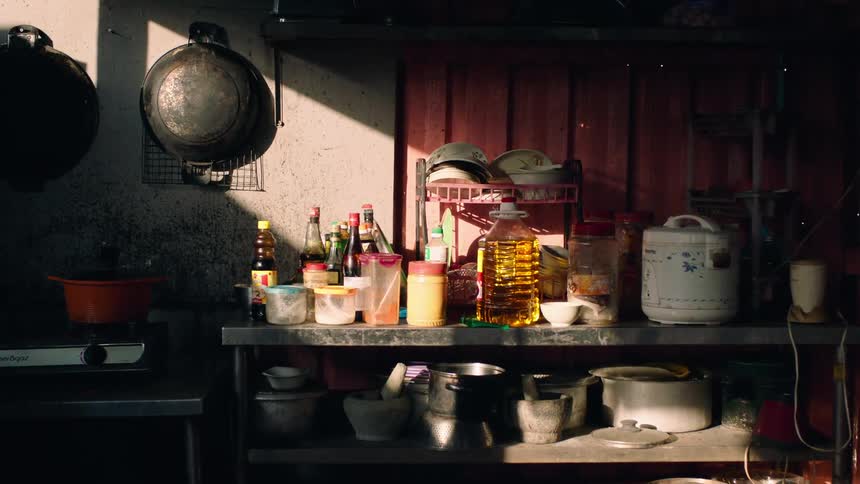 Sunlight illuminates cooking supplies on a shelf in a rustic outdoor kitchen as steam rises from a pot on the stove