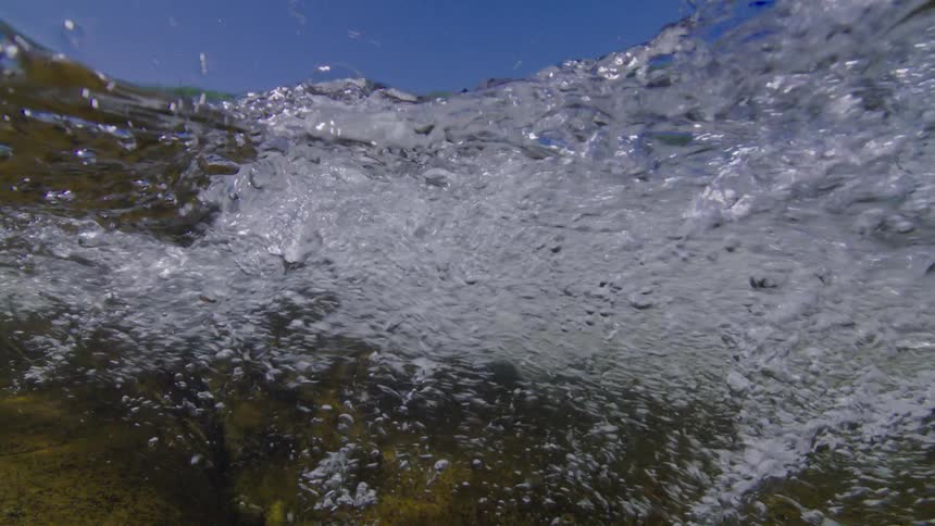 An immersive underwater perspective captures the turbulent, bubbling surface of a river with a split view of the blue sky and mountains above