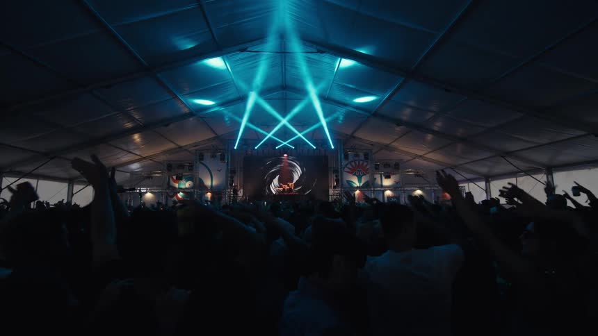 A massive crowd dances and cheers with hands in the air at a concert inside a large tent, illuminated by vibrant blue stage lights