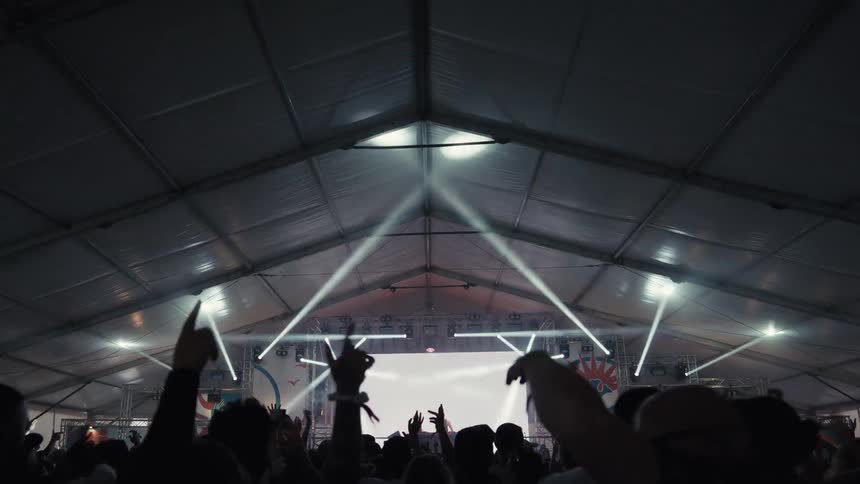 A vibrant crowd dances with hands in the air at a concert inside a festival tent with a dynamic stage and light show
