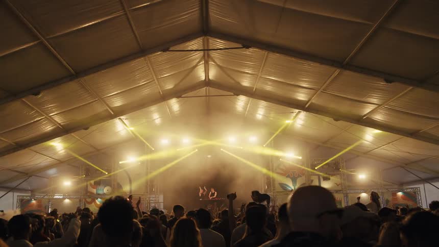 A massive crowd dances to a DJ set in a large festival tent with dramatic yellow stage lighting