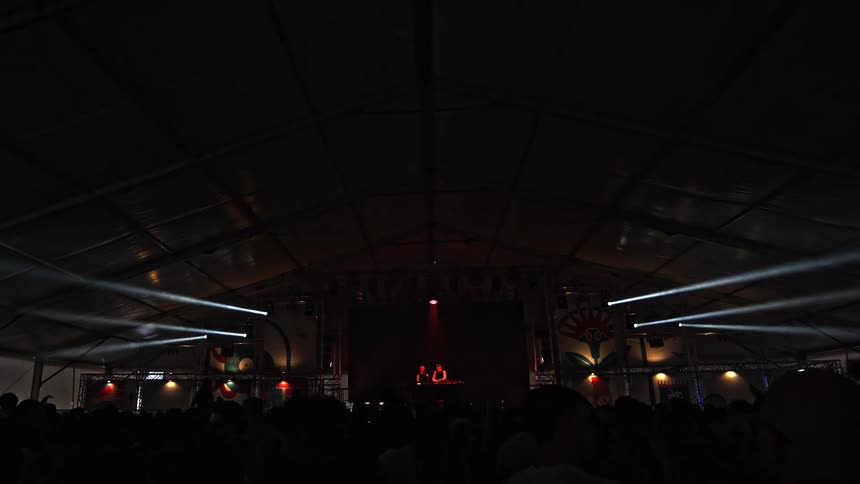 From the crowd's perspective, a DJ performs on stage with a vibrant light show and video screen inside a festival tent