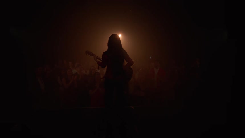 A silhouetted female musician walks onto a dark, hazy stage with her guitar to perform for a cheering crowd