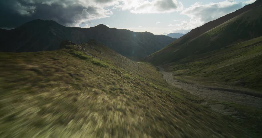 An exhilarating aerial flight along a rugged mountain ridge, descending into a vast, sunlit valley under a dramatic sky