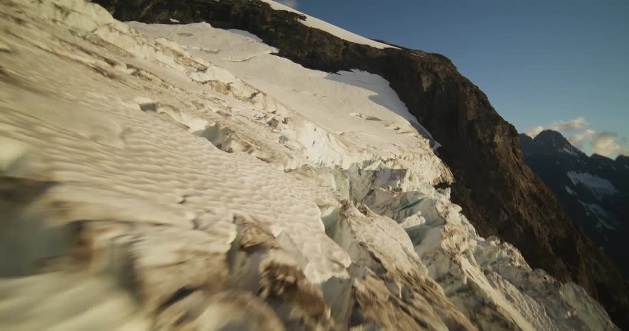 An immersive aerial journey backwards over the vast, crevassed expanse of a mountain glacier under a clear blue sky