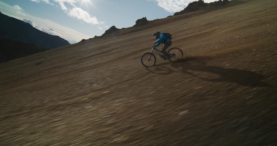 Epic aerial slow-motion shot of a mountain biker racing down a steep, loose scree slope in a vast mountain range