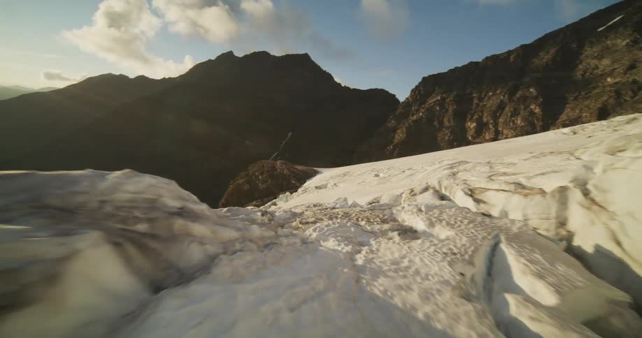 Smooth tracking shot glides over the textured surface of a mountain glacier, revealing a spectacular sunset over the valley