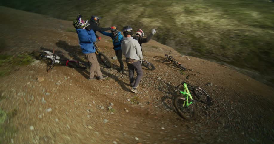 A dynamic circling shot captures a group of mountain bikers high-fiving and celebrating on a rugged alpine trail