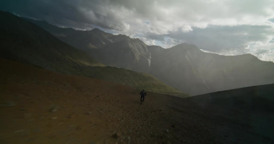 Slow motion drone shot follows an extreme mountain biker skillfully descending a steep, rugged mountain face under dramatic, sunlit clouds