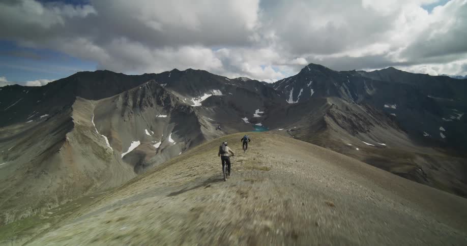 An epic aerial tracking shot follows two mountain bikers descending a steep, high-altitude ridgeline with a stunning turquoise alpine lake in the distance