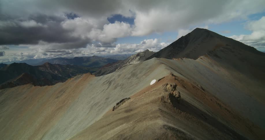 Soaring aerial flight over a barren mountain ridge reveals a vast, rugged landscape under a dramatic cloudy sky