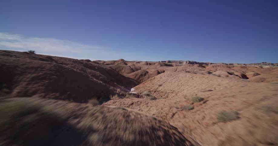 A first-person view drone soars at high speed through the winding, eroded canyons of a vast desert landscape