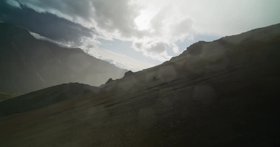 A lone mountain biker descends a steep and dusty mountainside in slow motion, silhouetted against a dramatic, backlit sky