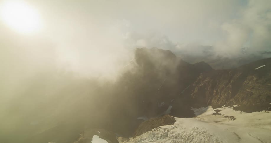 An ethereal aerial view pulls back through sunlit clouds to reveal a majestic, snow-capped mountain range