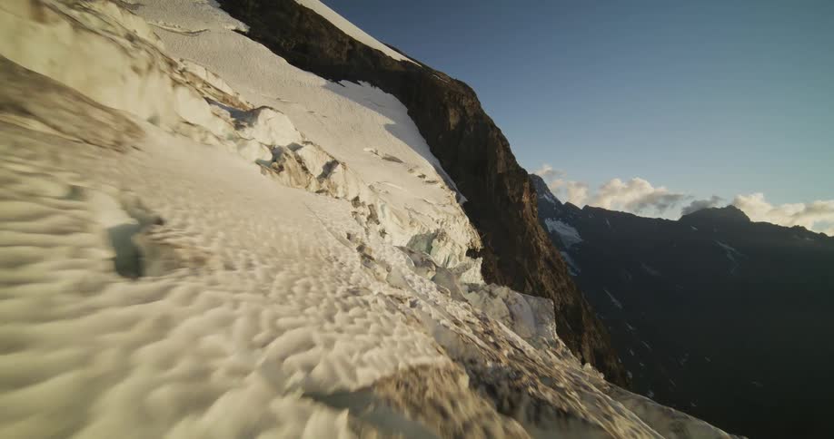 An epic aerial flight soars over the edge of a vast glacier, revealing a stunning mountain valley as the sun rises over the peaks
