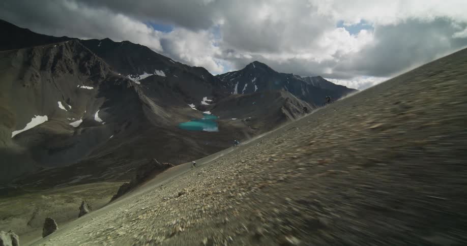 Aerial slow-motion view of three adventurous mountain bikers racing down a steep, rocky mountainside above a stunning turquoise alpine lake