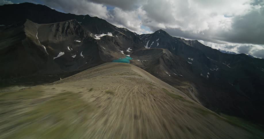 A dramatic aerial flight soars up a rugged mountain ridge, revealing a vast valley with a brilliant turquoise alpine lake