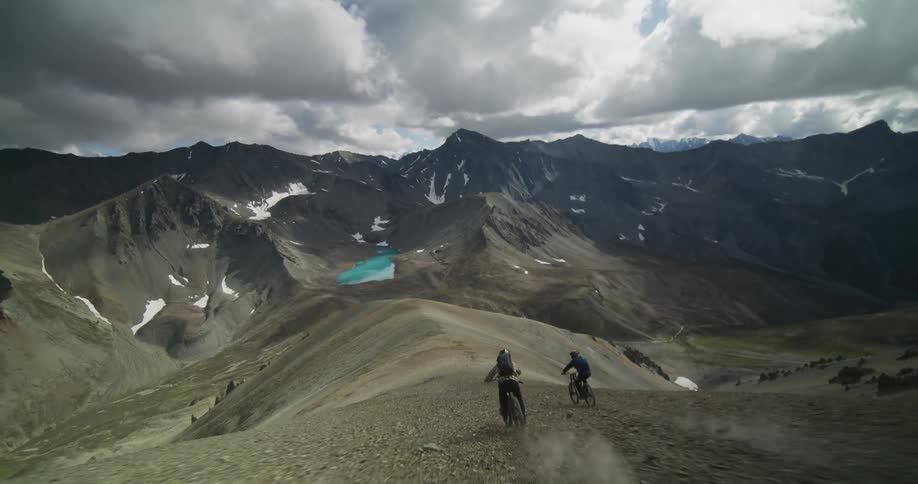 An exhilarating point-of-view shot follows two mountain bikers as they speed down a rocky mountain pass with an alpine lake in the distance