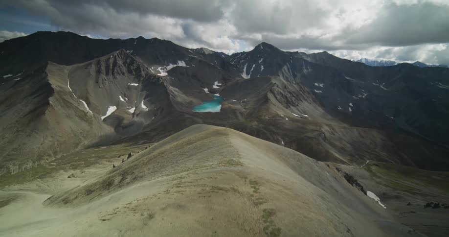 A stunning aerial view flies down a rocky ridge to reveal a vast mountain range and a brilliant turquoise alpine lake under a dramatic cloudy sky