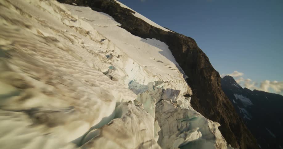 An aerial flight glides over the textured, snowy surface of a vast mountain glacier towards its icy, fractured edge
