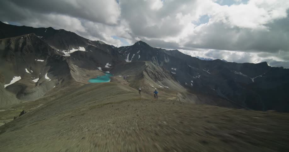An aerial view follows two mountain bikers riding along a high, rugged alpine ridge above a turquoise lake