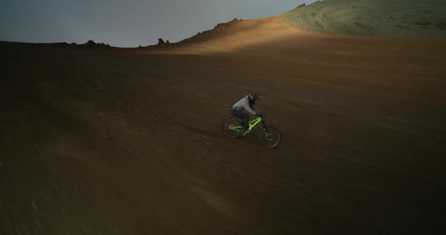 An aerial view follows an adventurous mountain biker as they race down a steep, barren mountainside in a vast and dramatic landscape