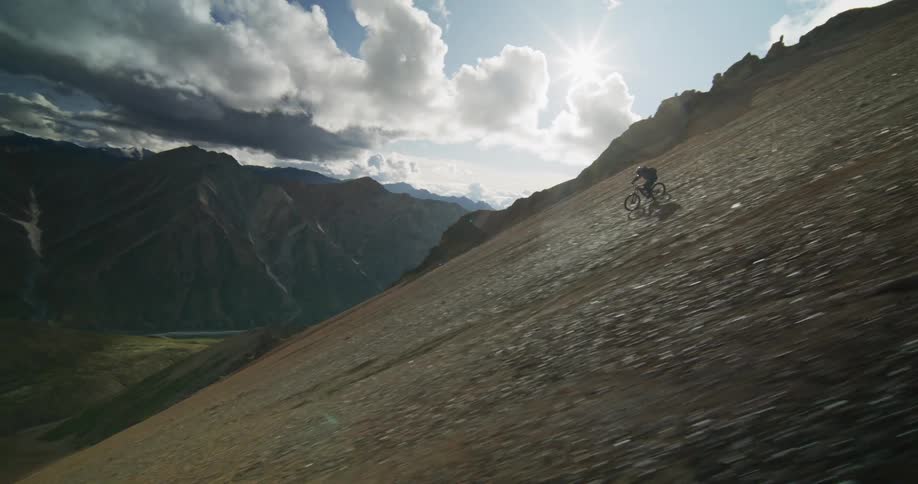 A lone mountain biker skillfully navigates a steep and challenging scree slope under a bright sun in a vast, epic mountain landscape