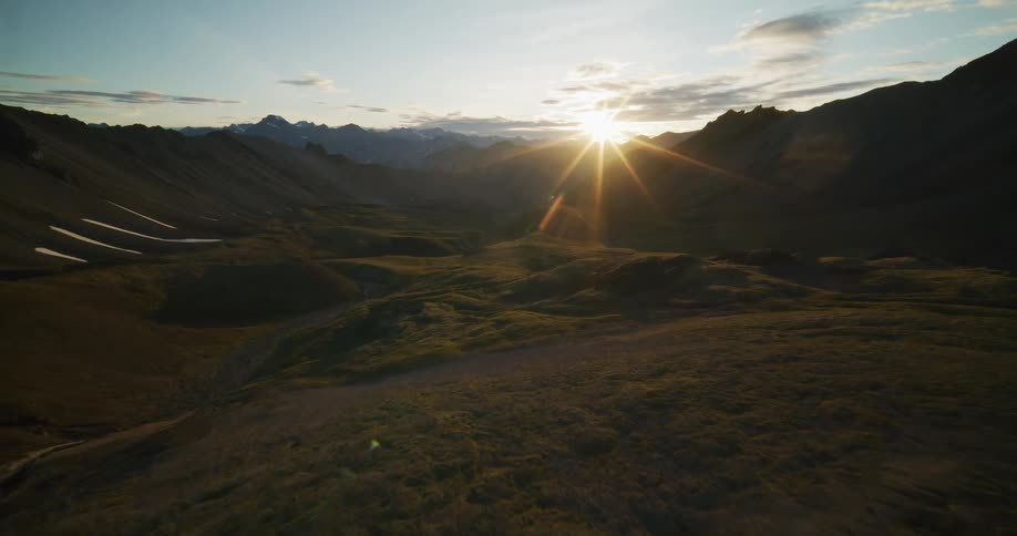 A thrilling FPV drone flight soars low through a vast, rugged mountain valley with patches of snow during a spectacular sunrise