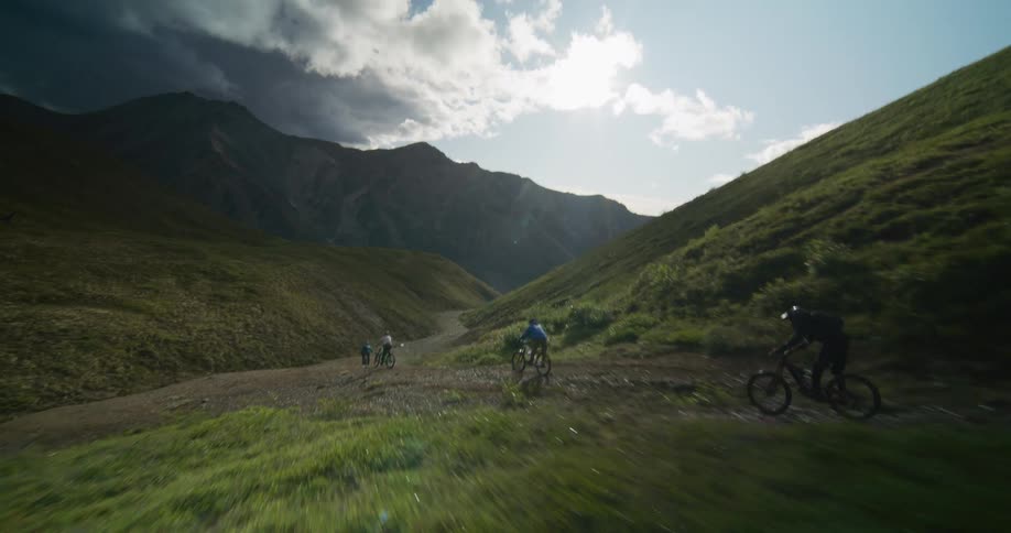 A group of mountain bikers descend a rugged trail through a vast, sun-drenched mountain valley