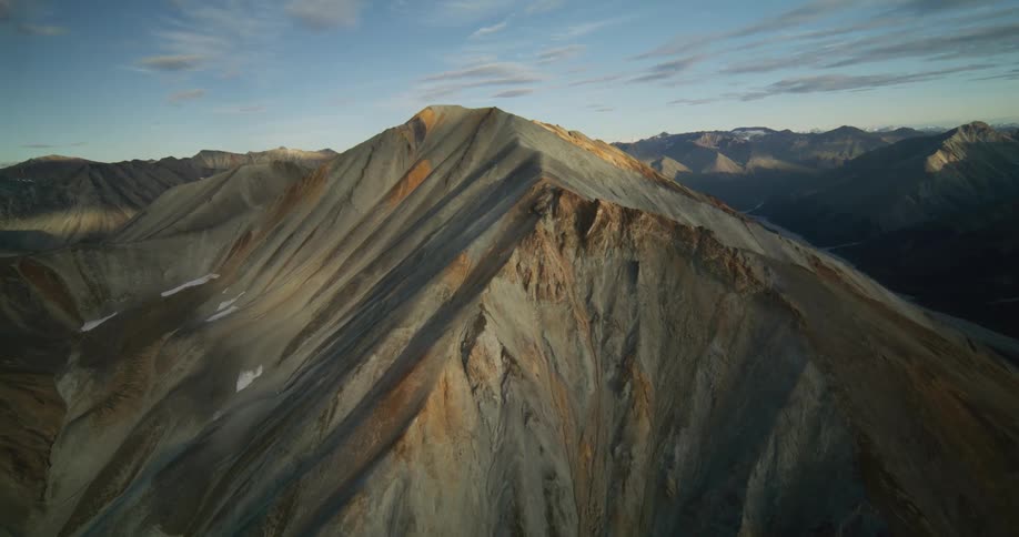 A static aerial shot shows a majestic, multicolored mountain peak with deep shadows cast by the rising sun
