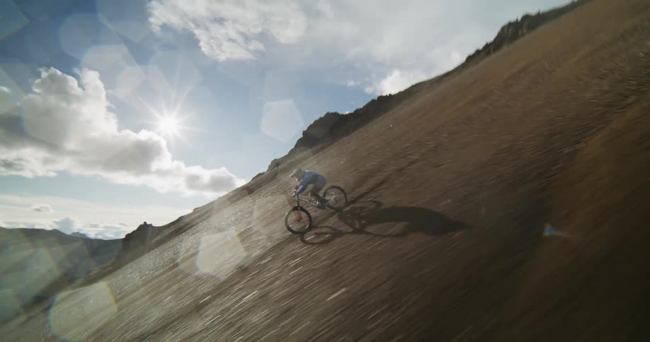 Dynamic slow-motion shot follows a professional mountain biker as he descends a very steep, loose scree slope in a vast, sunlit mountain range