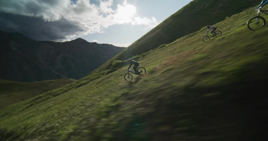 Dynamic slow-motion footage captures three mountain bikers riding down a steep, grassy mountain ridge against a dramatic sky