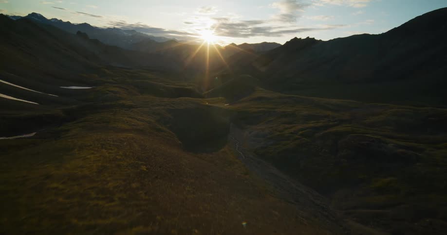 An aerial view flying through a vast mountain valley with patches of snow as the sun sets on the horizon