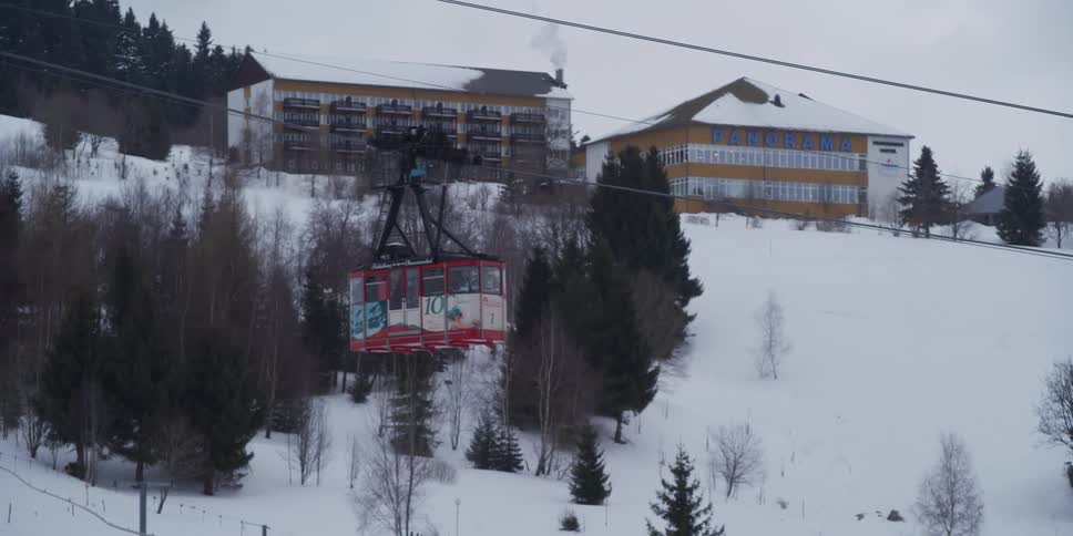 A red cable car gondola slowly travels up a snowy mountain past a ski resort on an overcast winter day
