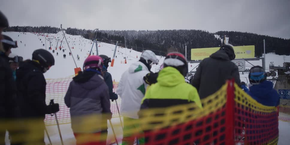 Skiers in colorful gear wait in line for the ski lift at a busy mountain resort on a snowy, overcast day