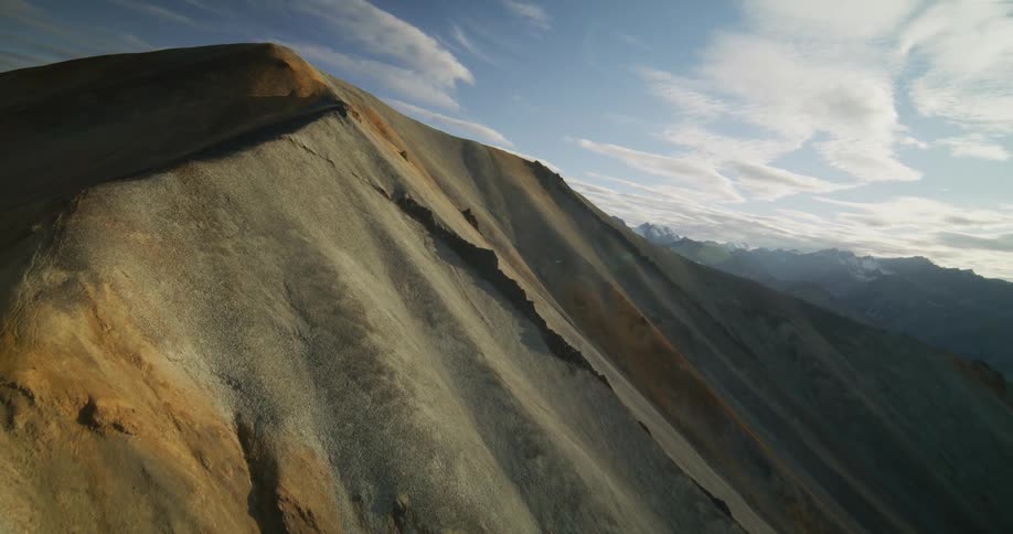 Aerial footage orbits a colorful, mineral-rich mountain ridge at sunrise, with a beautiful lens flare revealing the vast, epic landscape beyond