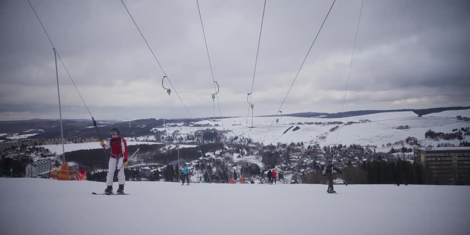 A female skier rides a surface lift up a snowy mountain slope with a beautiful view of a winter village below