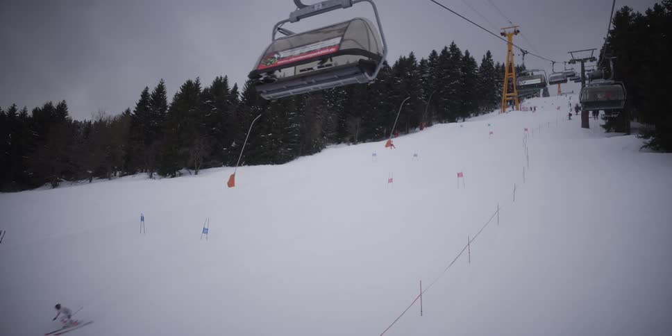 A skier races down a snowy slalom course, falling near a gate as a chairlift moves overhead on an overcast day
