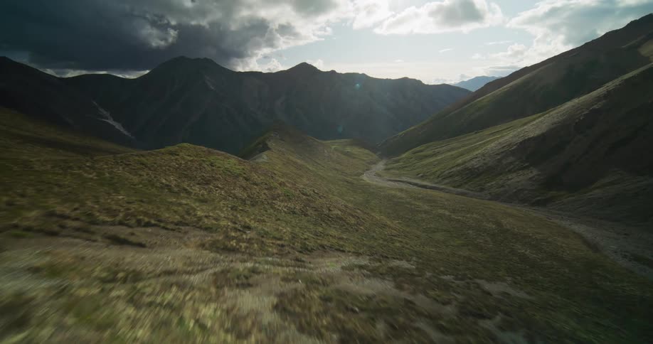 A dramatic aerial pullback shot flies low and fast over a vast green mountain valley with the sun shining through dark clouds