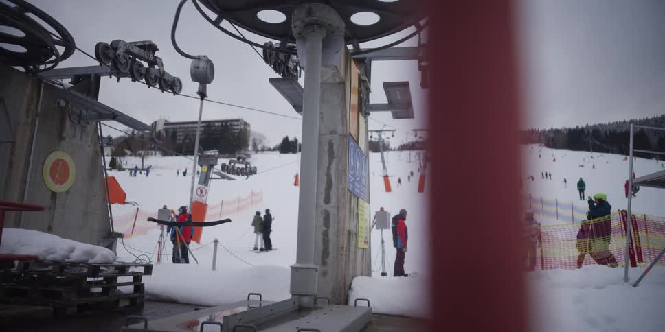 A T-bar ski lift operates at a busy ski resort, transporting skiers up the snowy mountain slope on a cloudy winter day