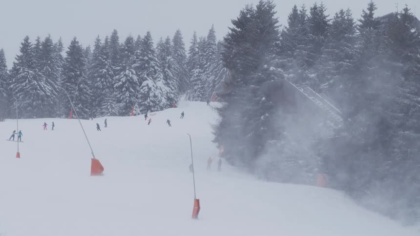 A snow cannon sprays fresh powder on a busy ski slope as people ski down the mountain through a snowy forest