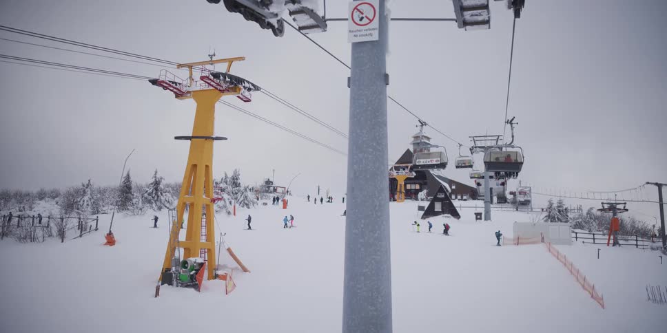 A point of view from a moving ski lift reveals the bustling slopes of a mountain resort on a snowy, overcast winter day