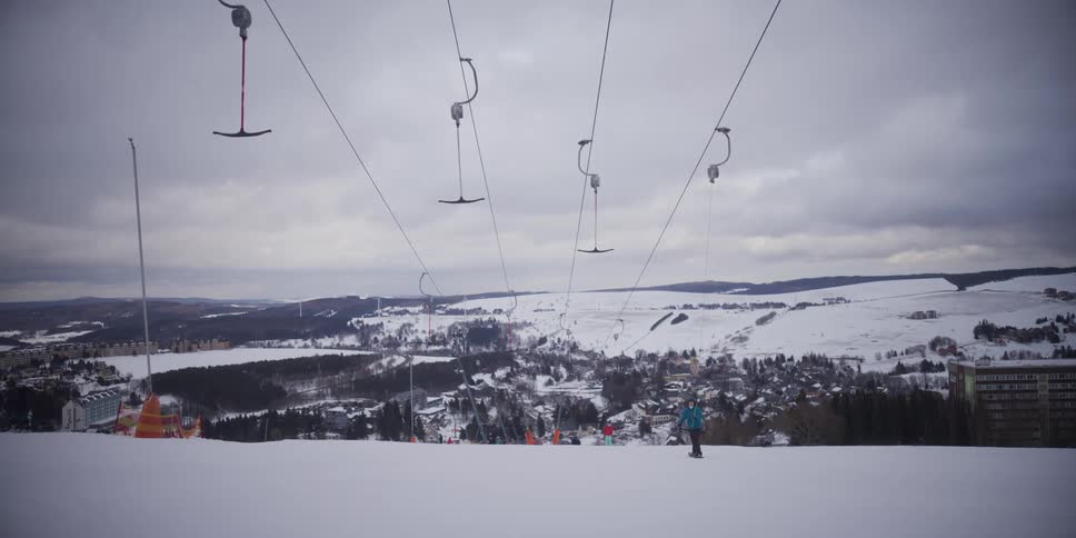 A lone snowboarder glides down a snowy slope at a ski resort with a T-bar lift overhead and a panoramic view of a snow-covered valley