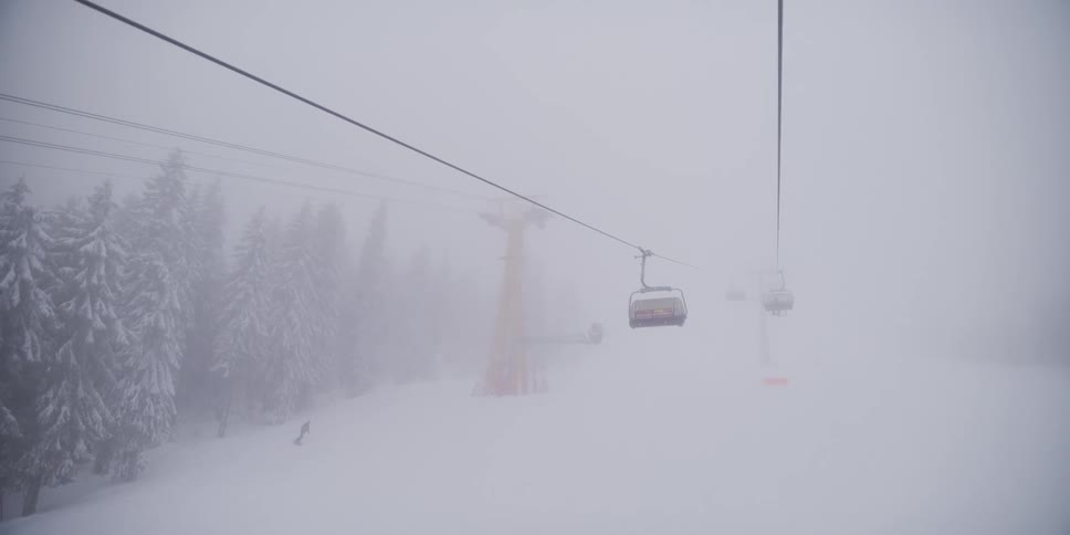 A view from a ski lift shows other chairs disappearing into the dense fog as a lone snowboarder carves down the snowy mountain slope below