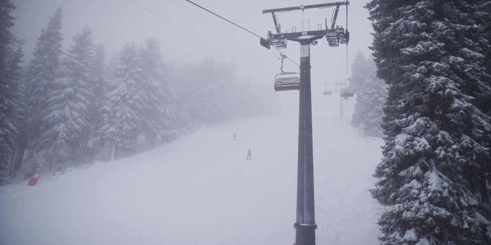 A point-of-view shot from a moving chairlift ascending a foggy, snowy mountain with skiers on the slope below